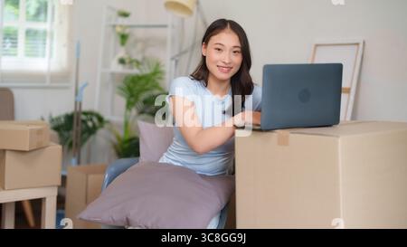 Femme adulte asiatique utilise heureusement la planification informatique de la nouvelle relocalisation d'appartement elle sourit gérant la logistique et la configuration de jour de déménagement à la maison Banque D'Images