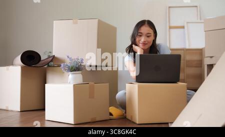 Jeune femme asiatique souriant adulte utilise un ordinateur à la maison entouré de boîtes d'emballage nouvelle propriété expérience de changement de style de vie Banque D'Images