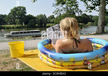 Jeune femme lisant un magazine dans une pataugeoire près du lac pour se rafraîchir sous la chaleur estivale (Aasee à Münster, Allemagne) Banque D'Images