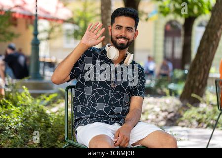 Joyeux jeune homme indien touriste souriant amical à la caméra, agitant les mains geste invitation bonjour, salut, salutation, accueillant avec expression hospitalière à l'extérieur. Guy est assis sur une chaise dans la rue de la ville. Banque D'Images