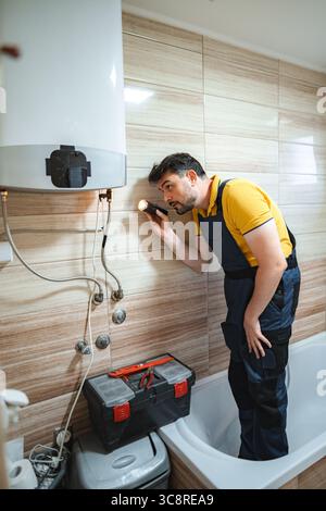 Plombier inspectant le chauffe-eau dans la salle de bains à l'aide de la lampe de poche Banque D'Images