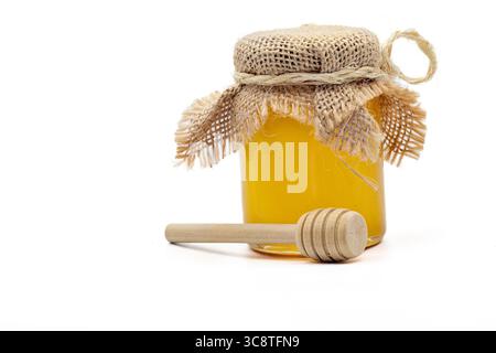 Bocal en verre rempli de miel doré, recouvert de toile de jute et de corde nouée, accompagné d'une benne à miel en bois sur fond blanc. Banque D'Images
