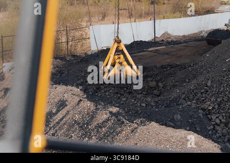 Un grand seau à charbon jaune planant au-dessus d'un monticule de charbon sur un site industriel, avec une grue excavatrice en fonctionnement. Le bras mécanique de la grue, Banque D'Images