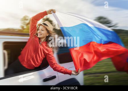 Une femme agite joyeusement le drapeau russe de la fenêtre d'une voiture en mouvement, célébrant la fête de la Russie. Vêtu d'une veste rouge éclatante et d'un chapeau élégant, sh Banque D'Images