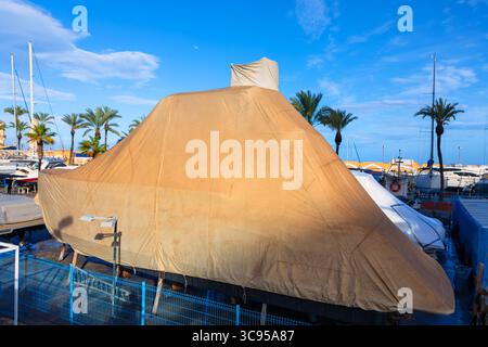 Yacht couvert en cale sèche, sécurisé sous une grande bâche de protection beige, entouré de palmiers et de bateaux amarrés sous un ciel bleu clair Banque D'Images