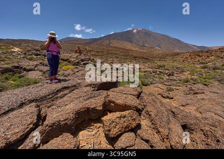 Randonneur féminin photographiant des paysages avec le stratovolcan du Teide à travers un champ de lave accidenté sous un ciel clair d'été Banque D'Images