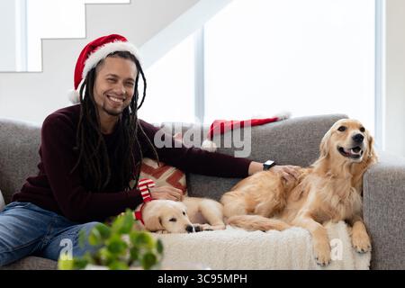 Homme portant chapeau de Père Noël et pull Bourgogne caressant Golden retrievers sur le canapé dans le salon Banque D'Images