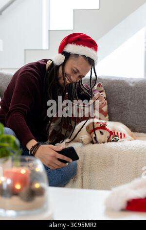 Homme afro-américain portant chapeau de Père Noël assis sur le canapé à la maison tenant le téléphone à côté du chien, terrarium Banque D'Images
