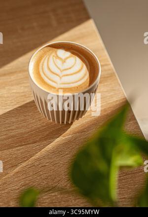 Une tasse de cappuccino en céramique agrémentée d'un élégant latte crème est posée sur une table en bois clair dans un café minimaliste. Banque D'Images