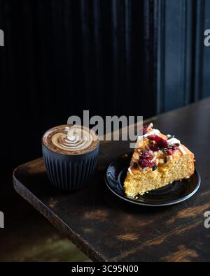 Une tranche de gâteau servie avec une tasse de cappuccino orné de latte art, placée sur une table rustique en bois. Scène de café confortable et accueillante. Banque D'Images