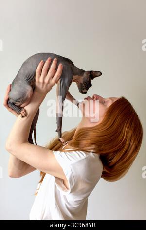 Une femme aux cheveux rouges soulève doucement son chat sphinx, partageant un moment tendre. Le fond Uni souligne leur lien et le style minimaliste. Le CA Banque D'Images