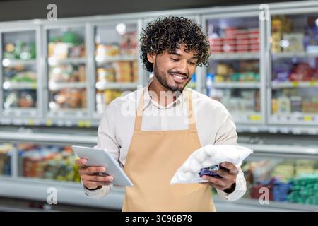 Un employé de supermarché regarde une tablette et un sac de produits surgelés, souriant tout en travaillant dans une épicerie. Banque D'Images