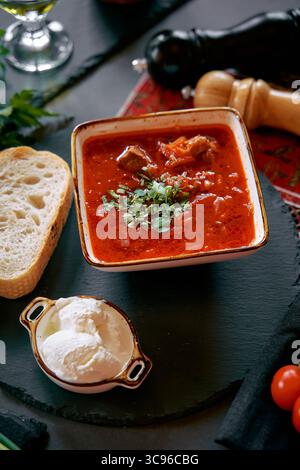 Un délicieux bol de soupe copieux à base de tomate avec viande tendre, herbes fraîches, garniture crémeuse et pain artisanal sur un plateau de service rustique Banque D'Images