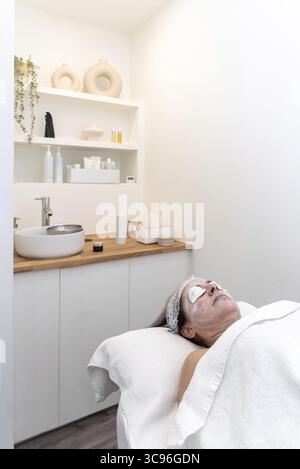 Femme senior bénéficiant d'un traitement du visage avec des coussinets pour les yeux et un masque facial, allongée sur une table de massage dans un salon de beauté moderne Banque D'Images