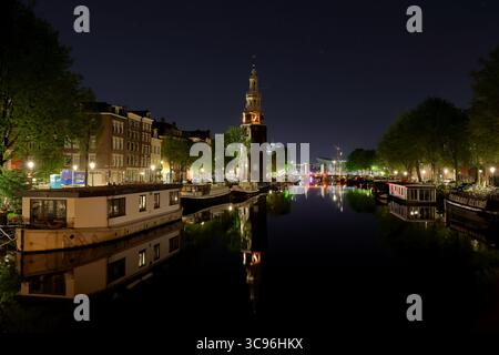 La tour Montelbaanstoren s'élève au-dessus du canal Oudeschans à Amsterdam tandis que les lumières chaudes scintillent sur l'eau, créant une scène nocturne sereine et fascinante. Banque D'Images