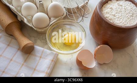 Oeufs frais dans un carton à côté d'une coquille fissurée et farine dans un bol, mis au four sur un comptoir de cuisine. Banque D'Images