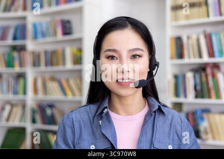 Une belle femme asiatique portant un casque sourit devant un fond d'étagère flou. Banque D'Images
