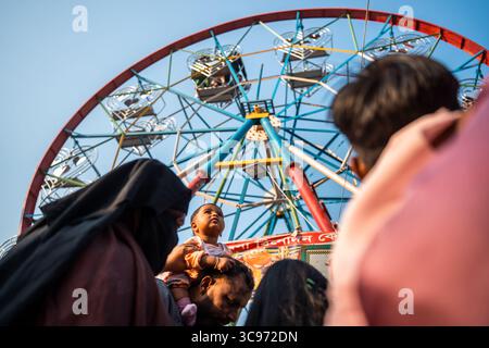 Bogura, Bangladesh - 03 novembre 2023 : vue sur la grande roue imposante contre le ciel dégagé, les enfants regardent vers le haut, portés sur les épaules, au milieu de l'atmosphère vibrante et animée du champ de foire. Banque D'Images