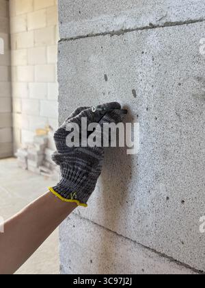 Une main dans un gant de protection tient un clou près d'un mur pour l'enfoncer. Chantier de construction. Rénovation, nouveau bâtiment. Banque D'Images