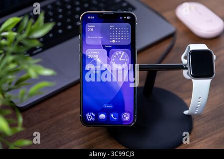 Poznan, Pologne - 29 juillet 2025 : ios 26 affiche des widgets dynamiques sur l'écran en verre liquide de l'iPhone d'Apple, se chargeant avec Apple Watch sur le bureau moderne Banque D'Images
