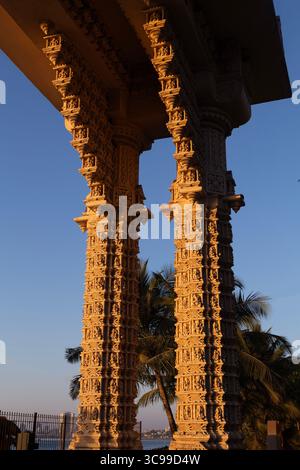 Des piliers en pierre finement sculptés avec des dessins traditionnels détaillés soutiennent un temple ou une structure de monument, assis contre un ciel bleu clair et des palmiers Banque D'Images