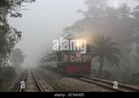 Chittagong, Bangladesh - 02 novembre 2018 : vue d'un train émergeant d'un épais brouillard, son front rouge et son corps jaune pâle coupant à travers les tons doux du paysage brumeux. Banque D'Images
