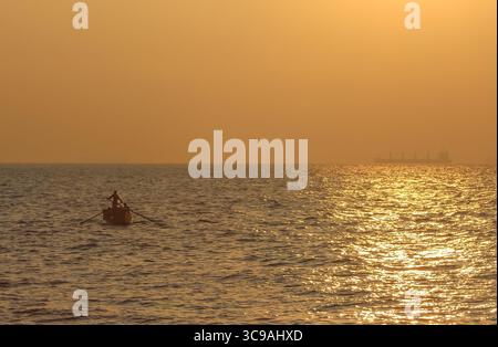 Chittagong, Bangladesh - 02 novembre 2018 : vue d'une figure solitaire ramant un petit bateau à travers l'eau chatoyante et ensoleillée, avec un navire lointain silhouette contre l'horizon doré. Banque D'Images