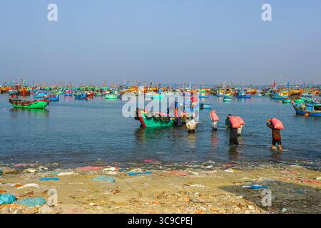 Mui ne, Vietnam-22 mars 2025 : pêcheurs transportant des sacs lourds au milieu de bateaux dynamiques dans le port de pêche Banque D'Images