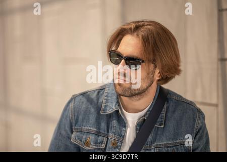 Un jeune homme élégant aux cheveux longs porte des lunettes de soleil, une veste en denim et un T-shirt blanc dans un portrait décontracté. Parfait pour la mode, le style de vie et vous Banque D'Images
