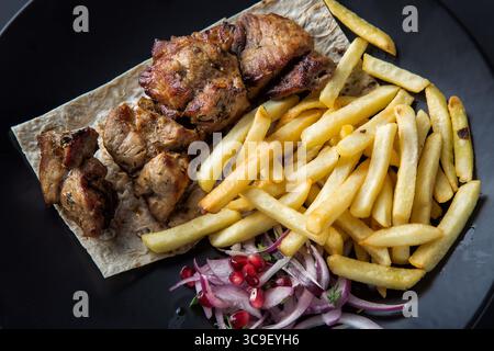 Shish kebab grillé servi avec frites, oignon et grains de grenade sur plaque noire de gros plan. Banque D'Images