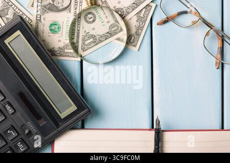 Concept commercial. Loupe avec lunettes, billets d'un dollar, cahier, stylo plume et calculatrice sur fond de bois bleu. Banque D'Images