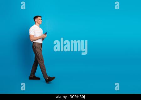 Jeune homme marchant sur fond bleu, tenant un smartphone, vêtu de façon décontractée d'un pantalon brun et d'un polo blanc, attitude joyeuse Banque D'Images