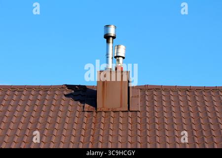 Deux cheminées modernes en métal s'élèvent d'un toit en tuiles brunes, nettement délimitées contre un ciel bleu sans nuages, montrant un mélange d'utilité et d'architecture Banque D'Images