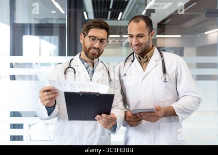 Deux médecins, en manteaux blancs avec stéthoscopes, examinent la paperasse et une tablette dans un cadre de bureau moderne. Ils semblent concentrés et collaboratifs. Banque D'Images