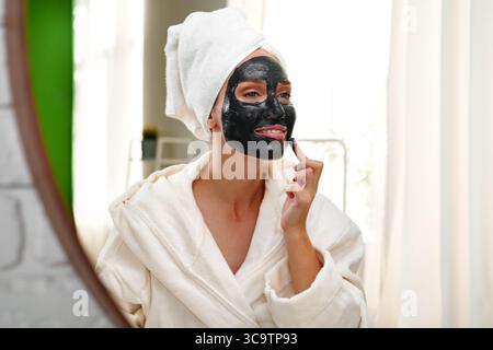 Femme appliquant un masque facial noir tout en regardant dans le miroir à la maison sur un après-midi de détente Banque D'Images