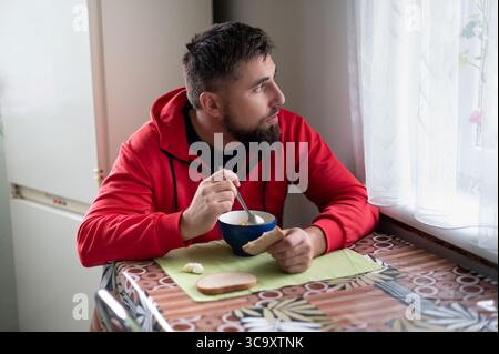 Cette scène capture un homme barbu dans un sweat à capuche rouge mangeant de la soupe à une table à motifs à côté d'une fenêtre avec des rideaux transparents, perdu dans ses pensées. Il détient bre Banque D'Images
