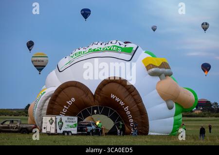 Chambley, France - 30 juillet 2025 : vue d'une montgolfière colossale, partiellement gonflée, ressemblant à une palette de peintre, entourée de montgolfières plus petites contre un ciel doux du soir. Banque D'Images