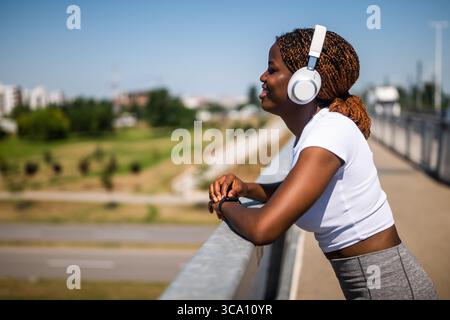 Fit femme sportive africaine écoutant de la musique avec des écouteurs tout en prenant une pause pendant son entraînement au pont de la ville. Banque D'Images