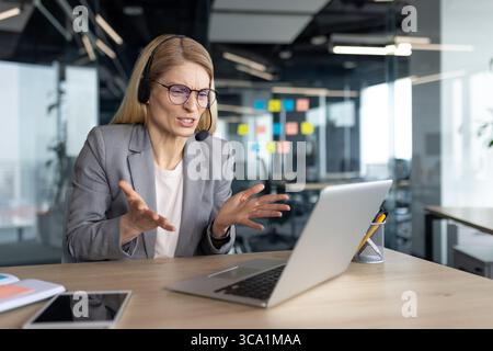 Femme nerveuse et en colère parlant tout en étant assise dans le bureau à l'aide d'un casque téléphone et ordinateur portable pour l'appel vidéo. Femme d'affaires en costume d'affaires à la conférence de réunion d'affaires en ligne. Banque D'Images