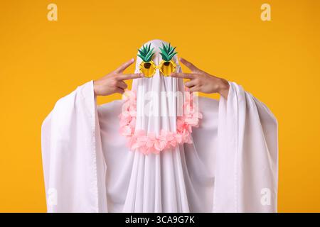 Fantôme drôle. Homme couvert de feuille blanche dans des lunettes de soleil et couronne de fleurs gestuelles sur fond orange Banque D'Images