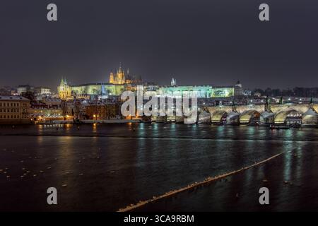 Illuminé le château de Prague et le pont Charles avec des toits enneigés reflétant la rivière Vltava par une nuit d'hiver calme. Banque D'Images