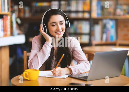Femme heureuse dans des écouteurs sans fil étudiant en ligne, en utilisant un ordinateur portable et en prenant des notes, assis au café, espace de copie Banque D'Images