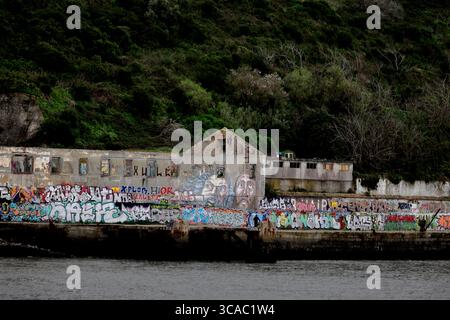Entrepôts riverains recouverts de graffitis à Praia das Lavadeiras, Almada, Lisbonne, au-dessous d'une colline verdoyante, capturant le caractère côtier urbain graveleux. Banque D'Images