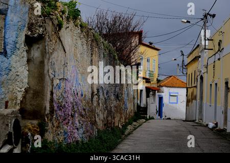 Ruelle étroite à Almada, Lisbonne, bordée par un mur strié de graffitis et des maisons pastel, lignes de services publics au-dessus, ciel couvert, usage éditorial. Banque D'Images