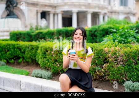 Jeune Latina joyeux avec des tresses assis sur un banc de jardin tenant une tasse de café. Porter un chemisier traditionnel coloré et une jupe dans le centre de Mexico Banque D'Images