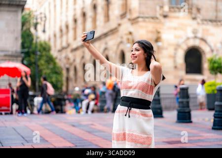 Heureux jeune Latina dans une robe corail et blanche tissée pose pour un selfie tout en se tenant près du Museo Nacional de Arte à Mexico, montrant fierté Banque D'Images