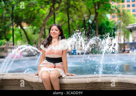 Jeune Latina heureuse dans une robe blanche et corail se trouve sur le bord d'une fontaine à Alameda Central, avec des jets d'eau et de la verdure autour d'elle dans le cœur Banque D'Images