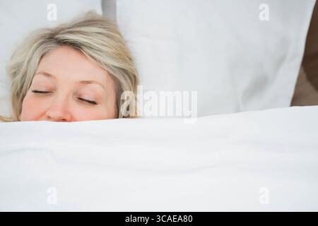 Femme mature couchée sous couette blanche dans la chambre avec cadre de lit, draps blancs et oreiller Banque D'Images
