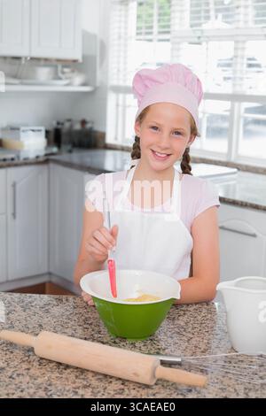 Enfant fille portant chapeau de chef rose et tablier remuant la pâte dans le bol de mélange dans la cuisine résidentielle Banque D'Images