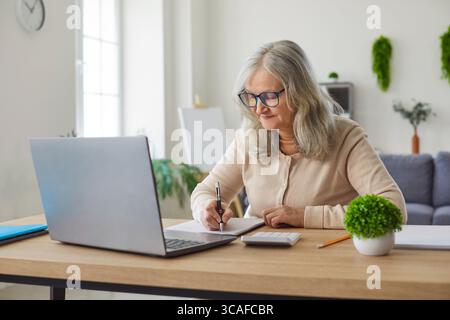 Femme âgée assise au bureau, à la maison, écriture plus ancienne dans le cahier, ordinateur portable, calculer le budget, apprendre Banque D'Images